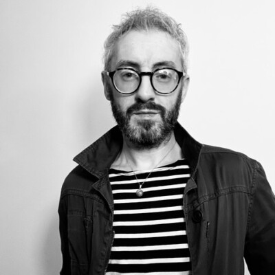 Black and white photo of Lorcan Black. He is a white man which short hair. He is wearing glasses, a dark jacket and a striped top.