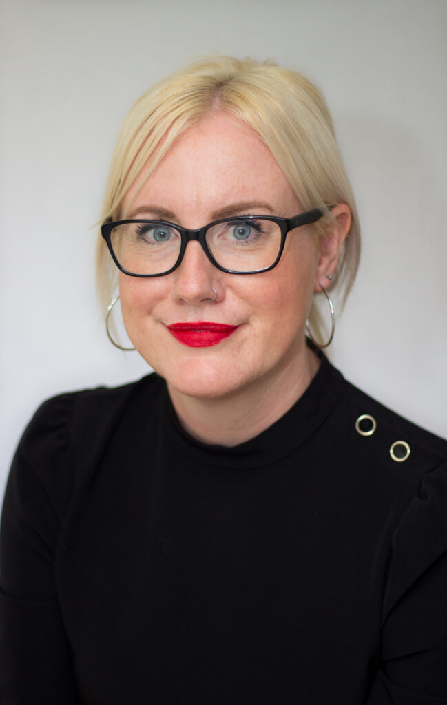 Wendy Allen is a white woman with short blonde hair. She is wearing black rimmed glasses, a black turtle neck and red lipstick.