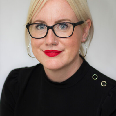 Wendy Allen is a white woman with short blonde hair. She is wearing black rimmed glasses, a black turtle neck and red lipstick.