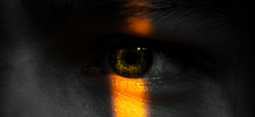 Close up photograph of a person's eye. The majority of their face is in darkness other than the iris which is illuminated by a vertical strip of light.