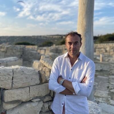 Joad R Wren is a white man with short dark hair. He is standing in front of an ancient greek ruin and is wearing a white linen shirt.