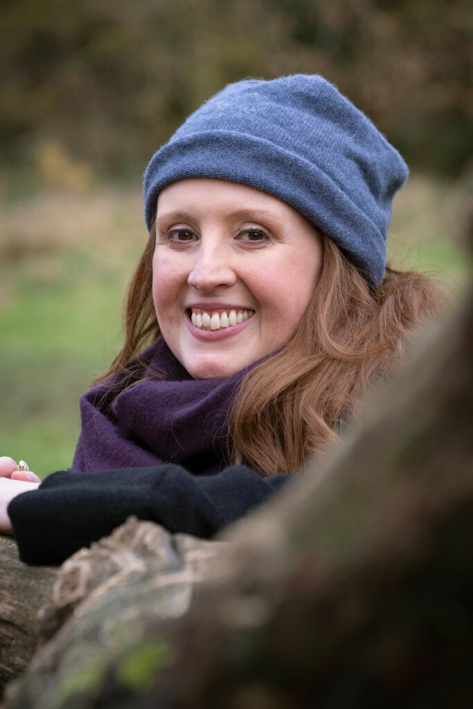 Kathryn Bevis is white woman with shoulder-length brown hair. She is wearing a blue beanie hat and a purple scarf and is looking at the camera and smiling.