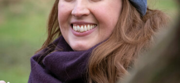 Kathryn Bevis is white woman with shoulder-length brown hair. She is wearing a blue beanie hat and a purple scarf and is looking at the camera and smiling.