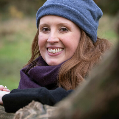 Kathryn Bevis is white woman with shoulder-length brown hair. She is wearing a blue beanie hat and a purple scarf and is looking at the camera and smiling.