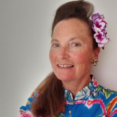Photo of Katrina Naomi wearing a blue floral top. Her hair is styled in a 1950s updo with pink flowers pinned above her left ear.
