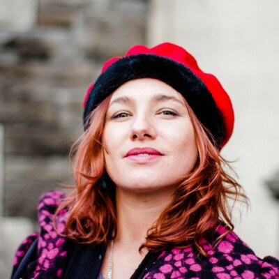 Gemma June Howell is a white woman with shoulder length auburn hair. She is wearing a red beret and a black and pink coat.