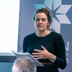 Photo of Bronwen Price speaking at an event. She has her hair up and is wearing a dark long-sleeved dress.