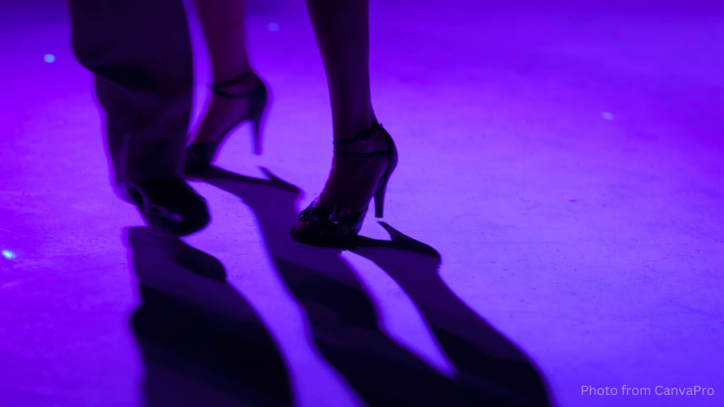 Close up of the feet of two dancers, their shadow stretching out on the purple-lit floor