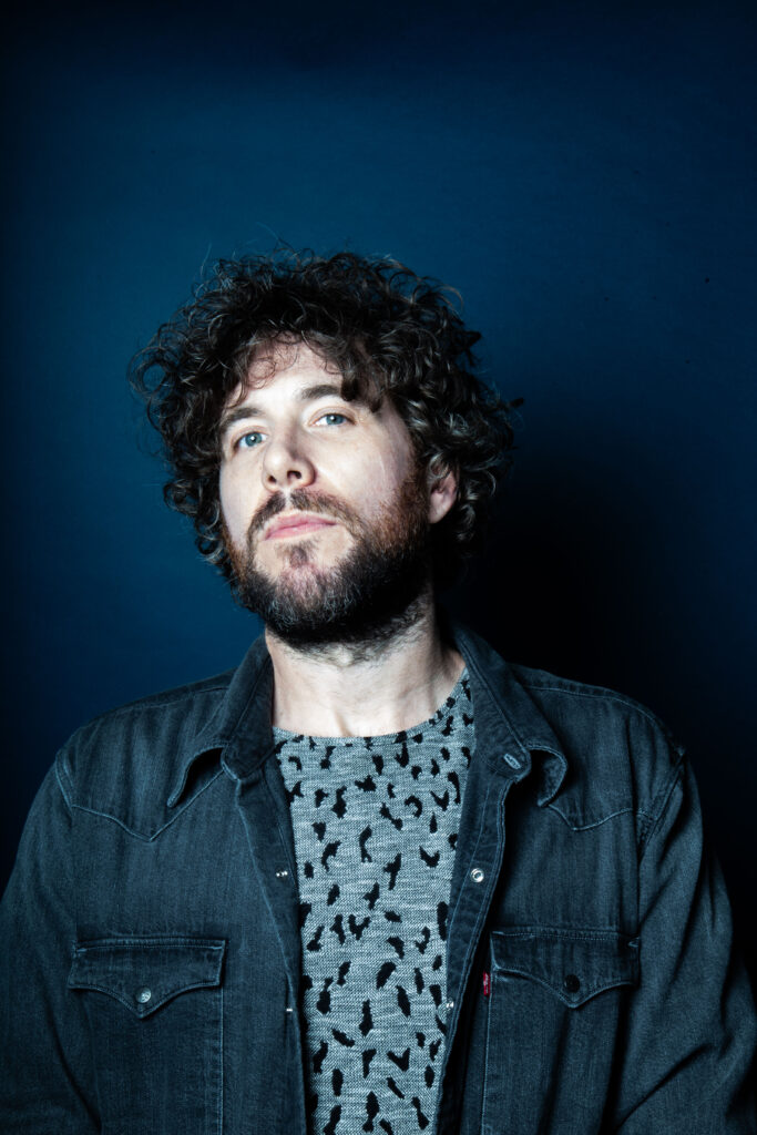 Photo of Jamie Morrison. He is standing against a dark blue background and is wearing a blue t-shirt and jacket. He has dark hair and a beard.