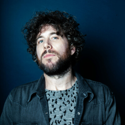 Photo of Jamie Morrison. He is standing against a dark blue background and is wearing a blue t-shirt and jacket. He has dark hair and a beard.