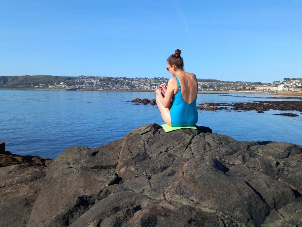 Katrina Naomi sitting on the rocks after a swim writing in the sunshine by the sea