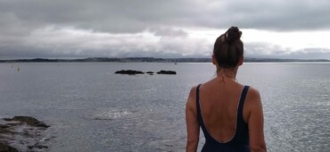 Katrina Naomi standing on the shore in her swimming costume looking out across the grey sea