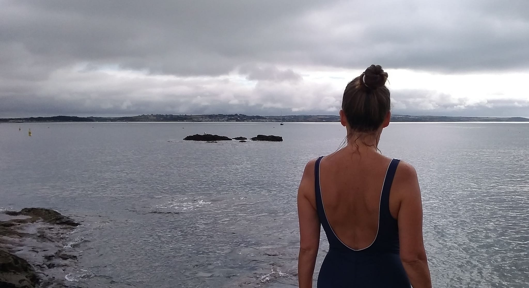 Katrina Naomi standing on the shore in her swimming costume looking out across the grey sea