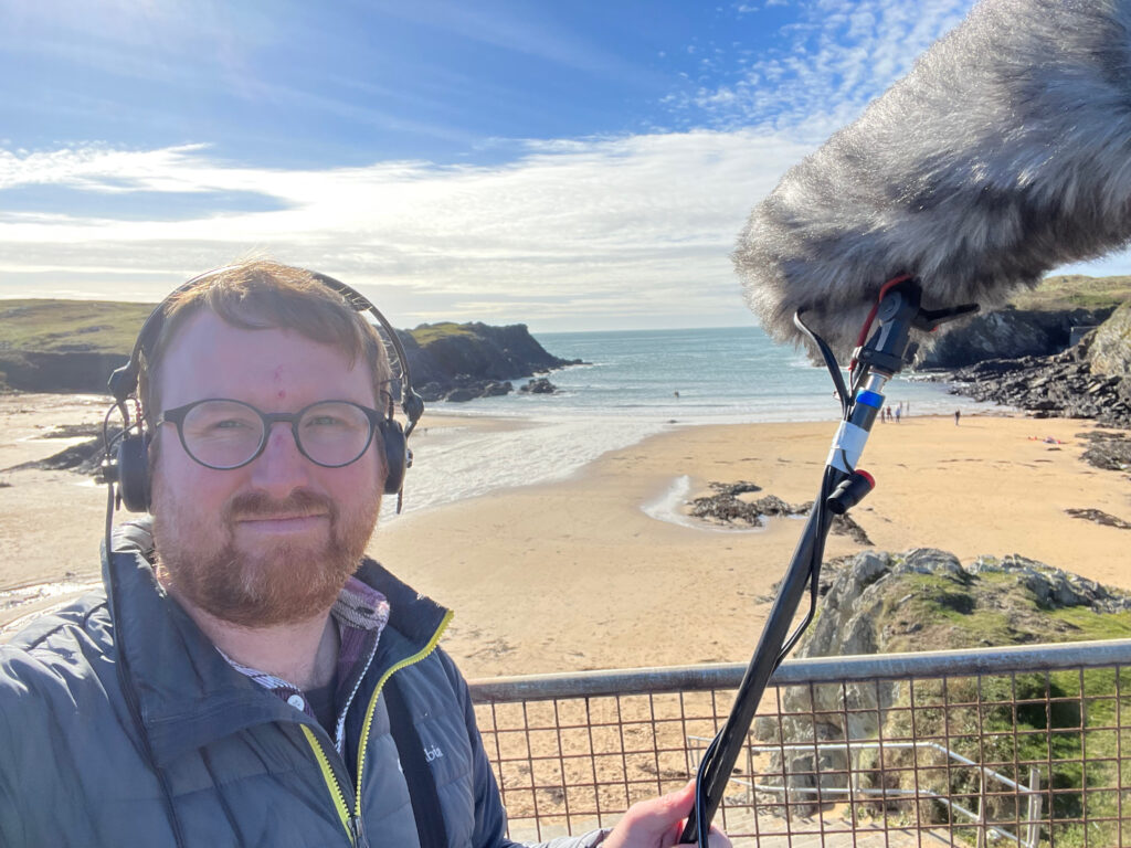 Photo of Meic Parry on the beach at Porth. He is holding a boom microphone and headphones.