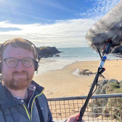 Photo of Meic Parry on the beach at Porth. He is holding a boom microphone and headphones.