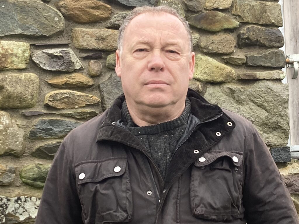 Photo of Sion Tecwyn standing in front of a stone wall. He is wearing a brown coat.