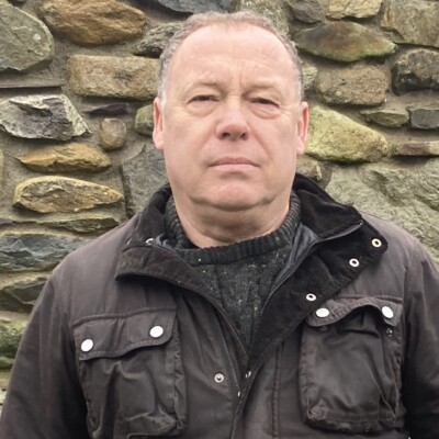 Photo of Sion Tecwyn standing in front of a stone wall. He is wearing a brown coat.