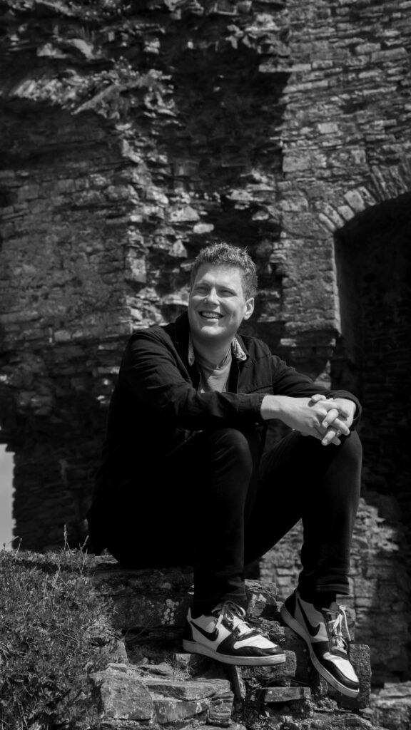 Black and white photo of Alex Hubbard sat on the walls of Aberystwyth castle.