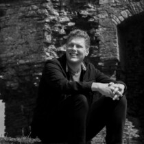 Black and white photo of Alex Hubbard sat on the walls of Aberystwyth castle.