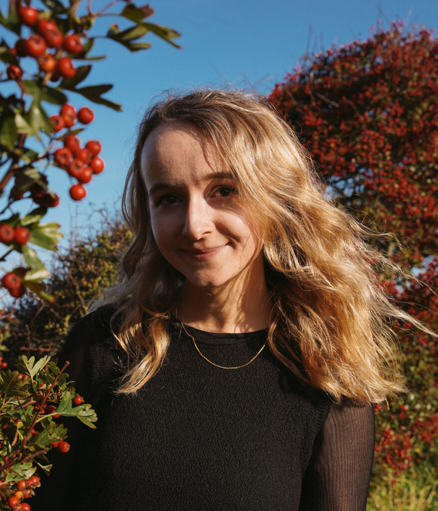 Colour photo of Efa Lois. She is outside by a colourful tree with red and green leaves and is wearing a black t-shirt.