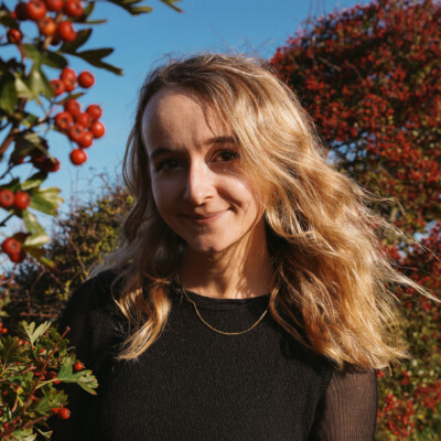 Colour photo of Efa Lois. She is outside by a colourful tree with red and green leaves and is wearing a black t-shirt.