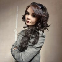Colour photograph of Natalie Ann Holborow. She is looking purposefully into the camera with her arms crossed and is wearing a grey blazer.