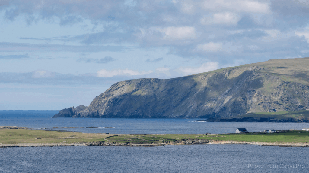 Photo of a green strip of land surrounded by sea with dramatic cliffs in the background.