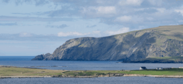 Photo of a green strip of land surrounded by sea with dramatic cliffs in the background.