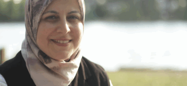 Photo of Abeer Ameer sitting outside by a lake. She is wearing a white shirt and patterned headscarf.