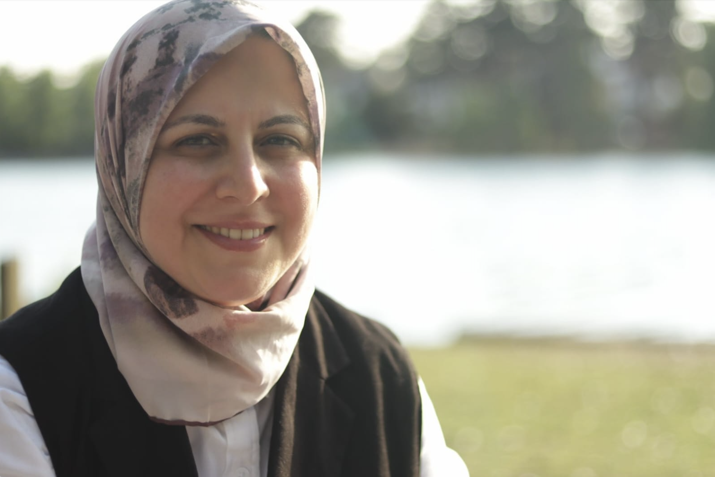 Photo of Abeer Ameer sitting outside by a lake. She is wearing a white shirt and patterned headscarf.