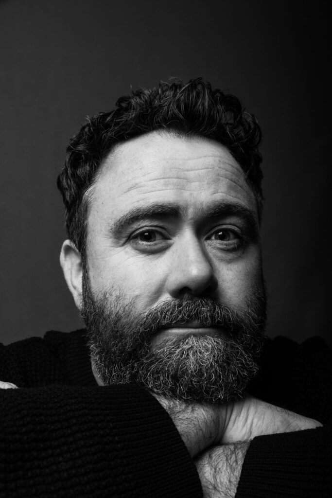 Black and white photo of Celyn Jones. He is resting his chin on top of his folded hands and has dark curly hair and a beard.