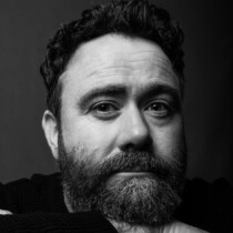 Black and white photo of Celyn Jones. He is resting his chin on top of his folded hands and has dark curly hair and a beard.