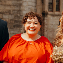 Colour photograph of Frances Turpin. She is standing outside in a bright orange dress and is smiling at the camera. She has dark curly hair.