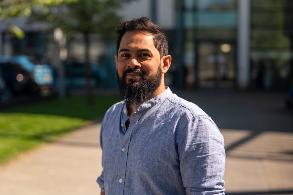 Photo of Azim Ahmed standing outside in the sunshine. He is wearing a blue shirt and has dark hair and a beard.