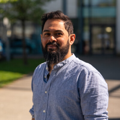 Photo of Azim Ahmed standing outside in the sunshine. He is wearing a blue shirt and has dark hair and a beard.