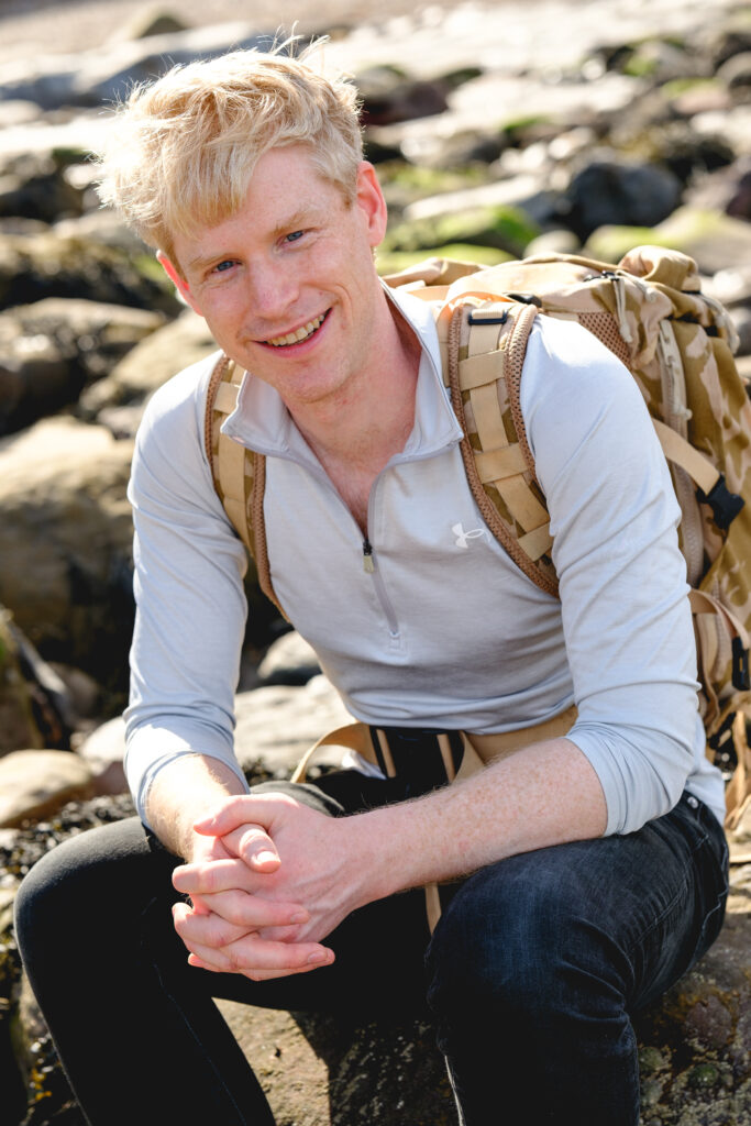 Photo of Matthew Myerscough sitting amongst rocks on the beach. He's wearing a pale fleece and a rucksack.