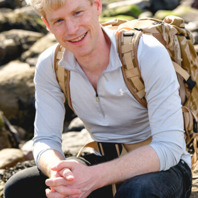 Photo of Matthew Myerscough sitting amongst rocks on the beach. He's wearing a pale fleece and a rucksack.