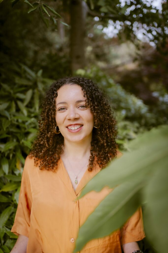 Photo of Taylor Edmonds. She is standing outside and is wearing a bright orange top.