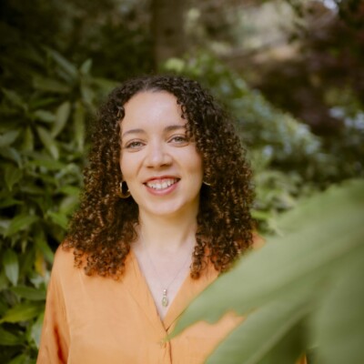 Photo of Taylor Edmonds. She is standing outside and is wearing a bright orange top.