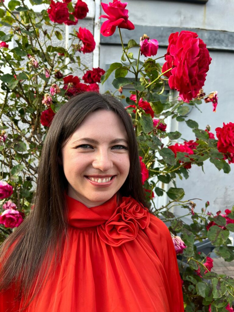 Photo of Alice Hughes stood outside in front of a blooming rose which matches her red blouse.