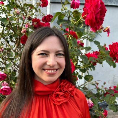 Photo of Alice Hughes stood outside in front of a blooming rose which matches her red blouse.