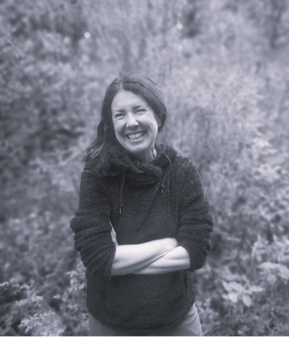 Black and white photo of Julie Brominicks standing outside. She is smiling at the camera with her arms crossed.